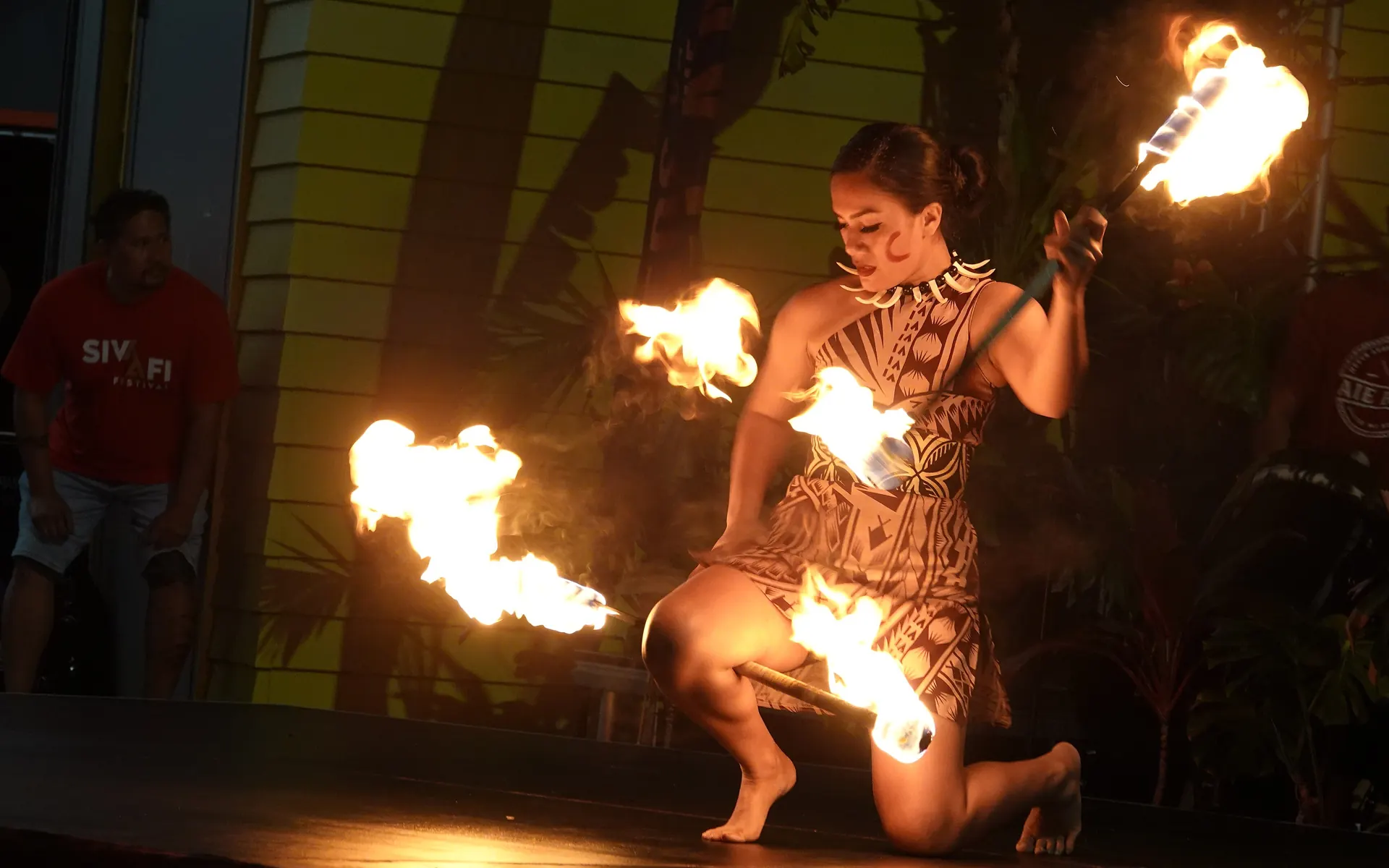 A Samoan fire dancer kneeling on a dark stage as two blazing fire knives spin around her, with tropical plants and a drumming band behind, photographed at a fire knife festival in Auckland